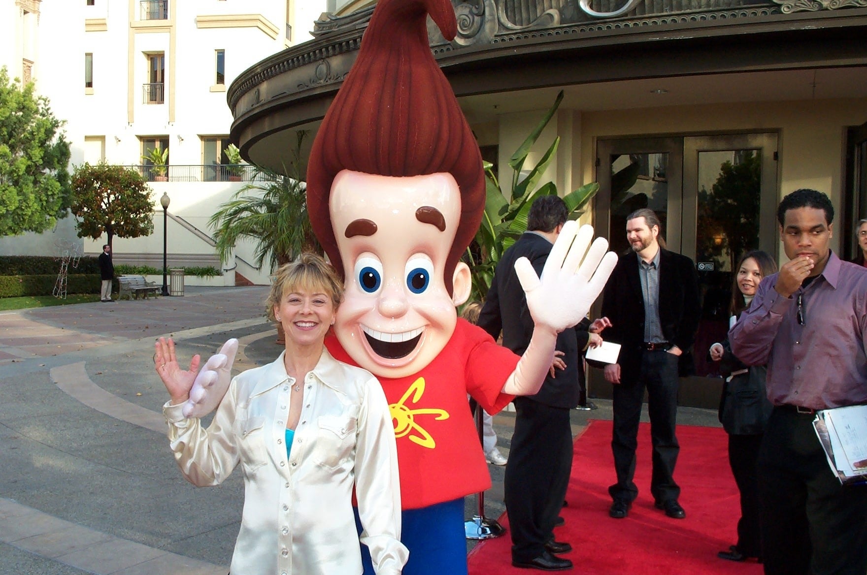 A woman in a satin blouse poses with someone in a Jimmy Neutron costume on a red carpet. Others are in the background