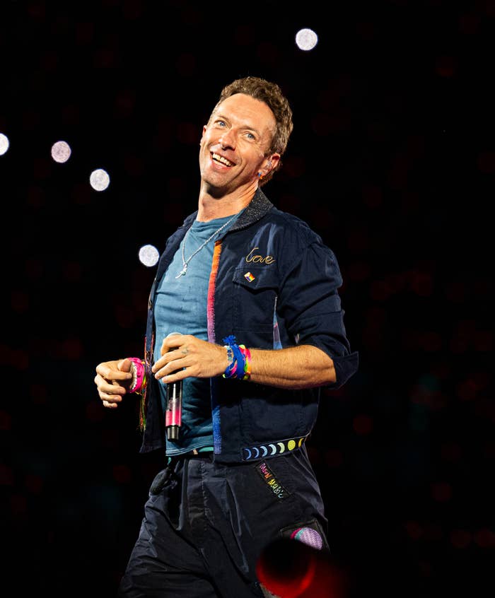 Person in colorful casual outfit holds microphone, smiling on stage during a performance, with bright background lights