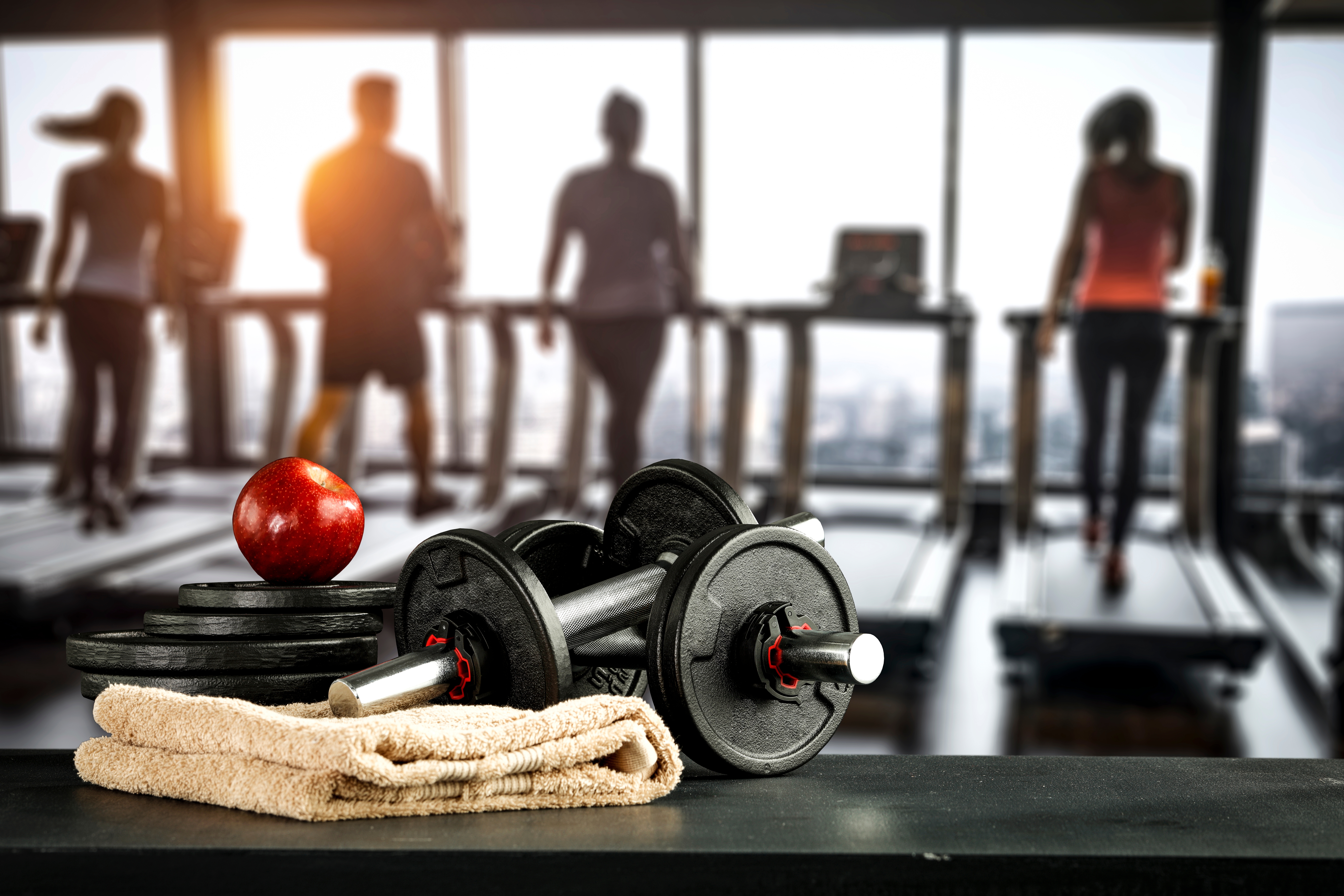 Dumbbells, weights, a towel, and an apple sit on a gym bench, while four people use treadmills in the background
