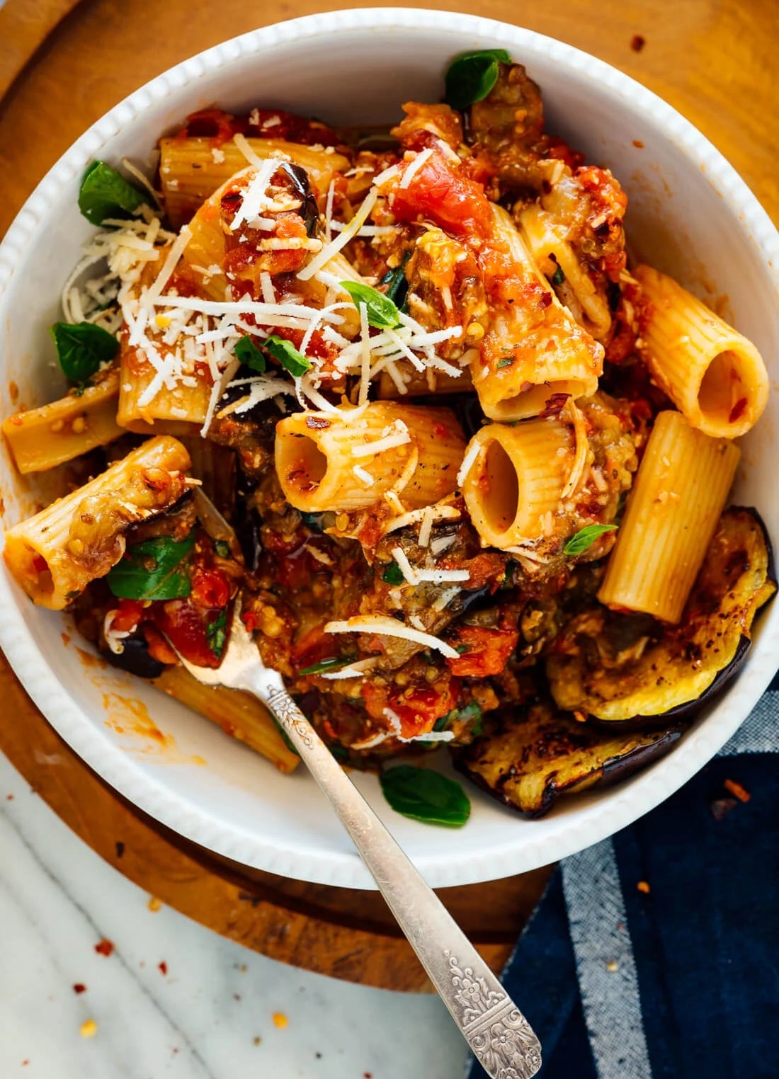 Plate of rigatoni pasta with herb  sauce, grated cheese, eggplant, and caller  basil leaves connected  a woody  board