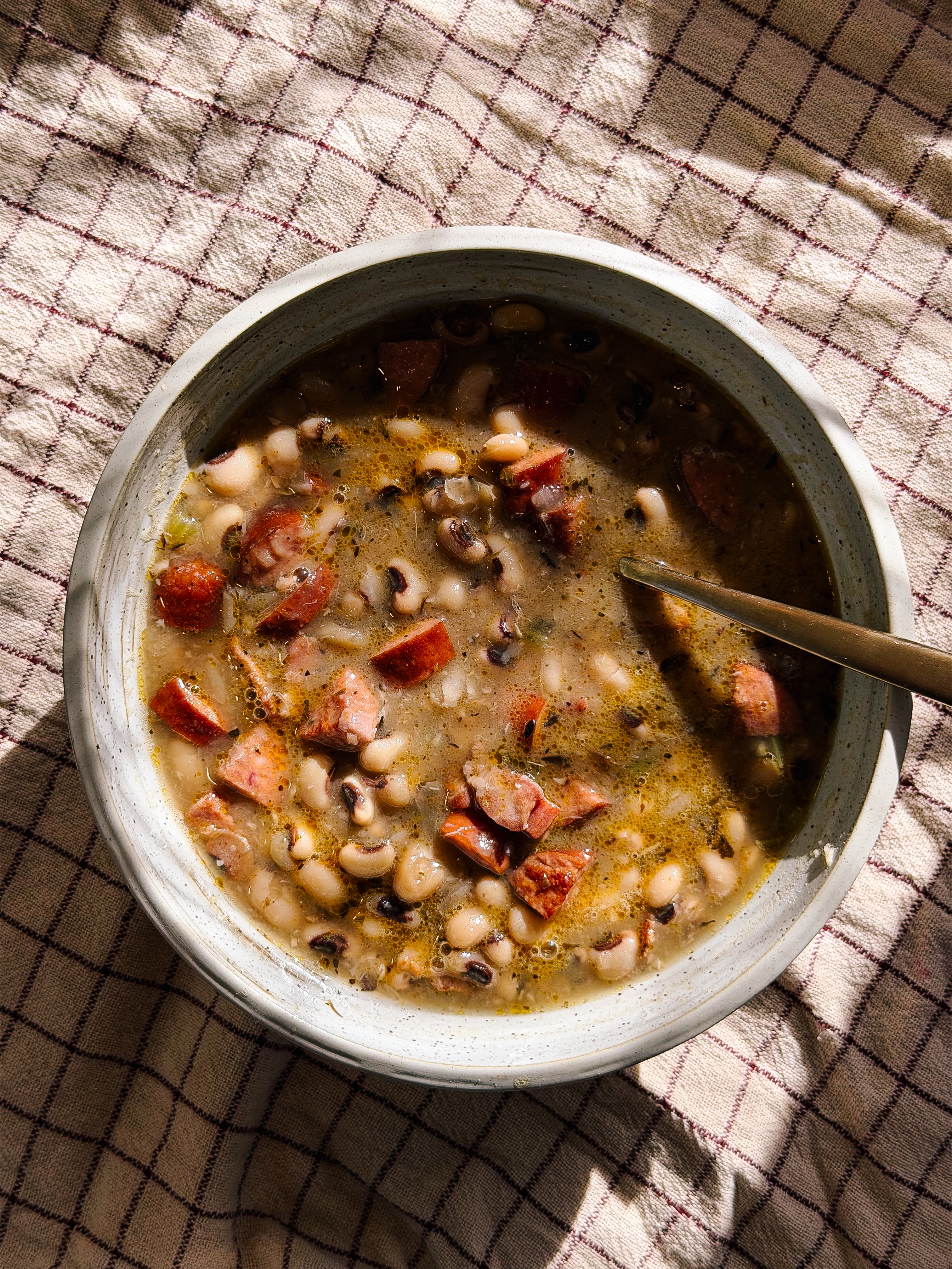 A vessel  of hearty legume  crockery   with chunks of meat, a spoon nestled inside, placed connected  a checked cloth surface