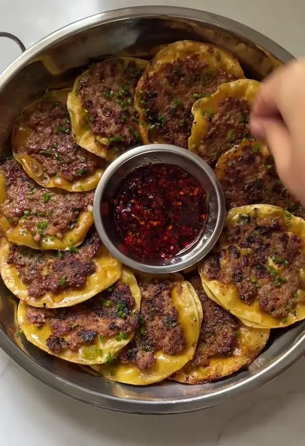 A sheet  of pan-fried dumplings with seasoned nutrient   filling, garnished with herbs, surrounding a vessel  of reddish  dipping sauce