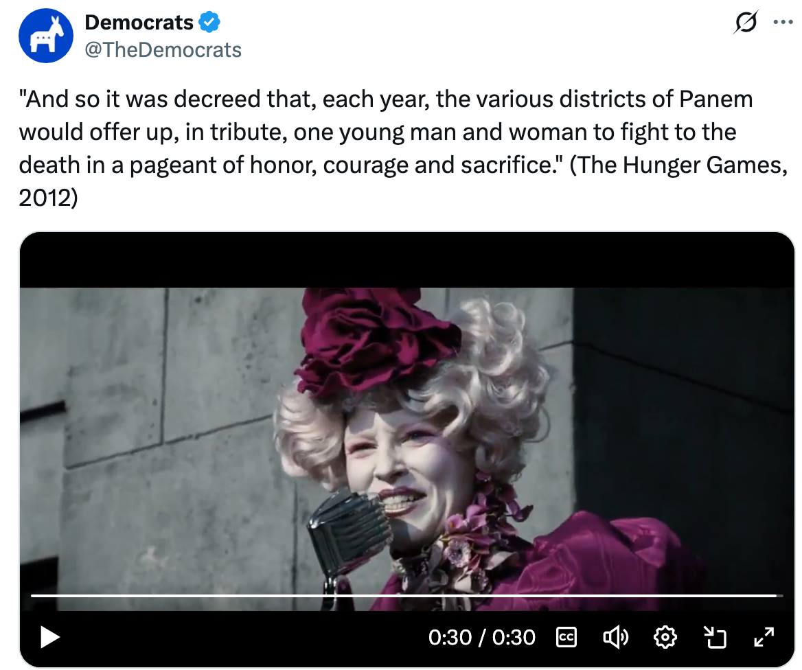 En person med forseggjort, teatralsk antrekk og frisyre snakker inn i en vintagemikrofon. Teksten refererer til "The Hunger Games" film sitat