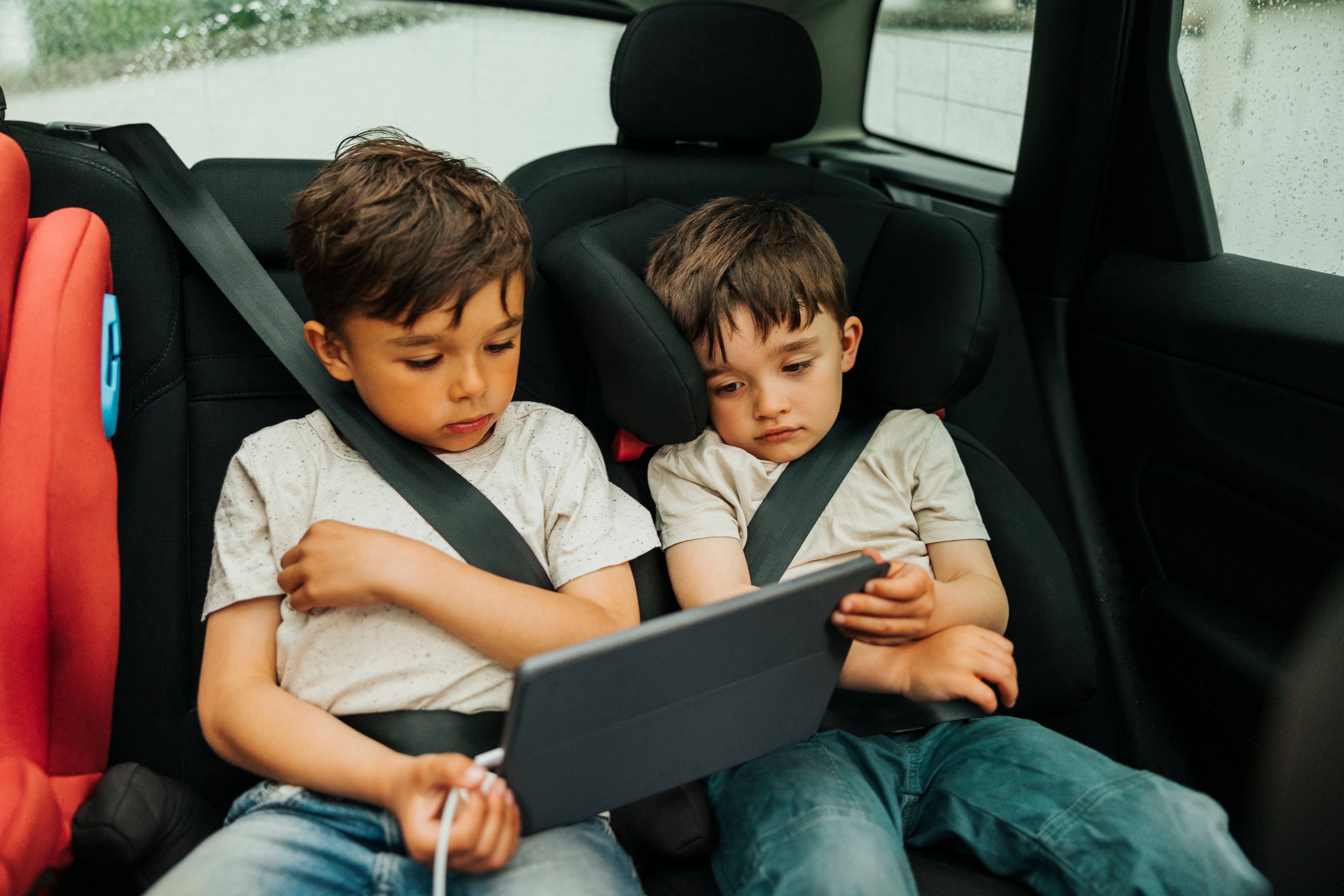 Two young children in car seats watch a tablet together in the backseat of a car