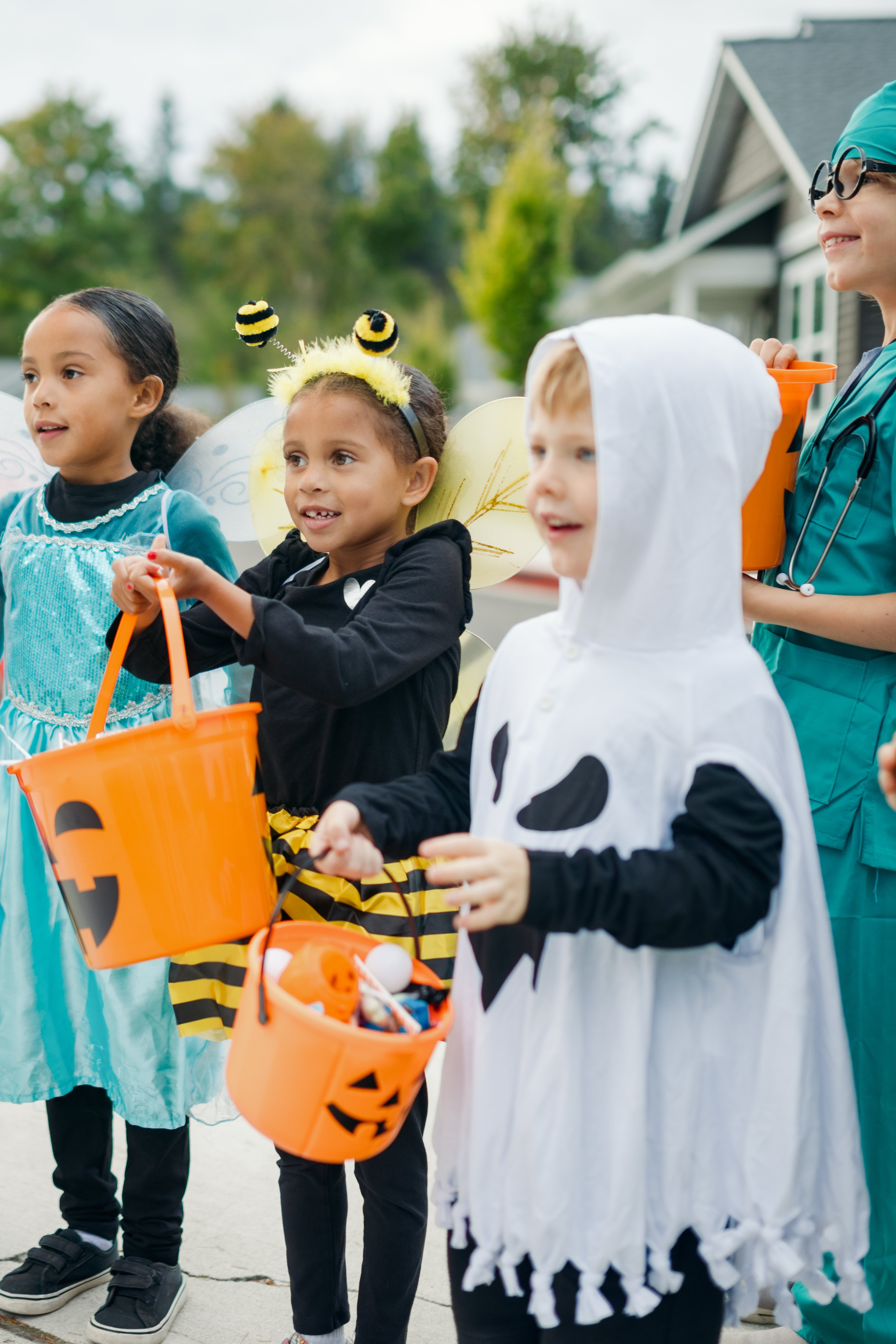 Children in Halloween costumes, including a bee, ghost, and princess, hold treat buckets while a smiling adult in scrubs watches them