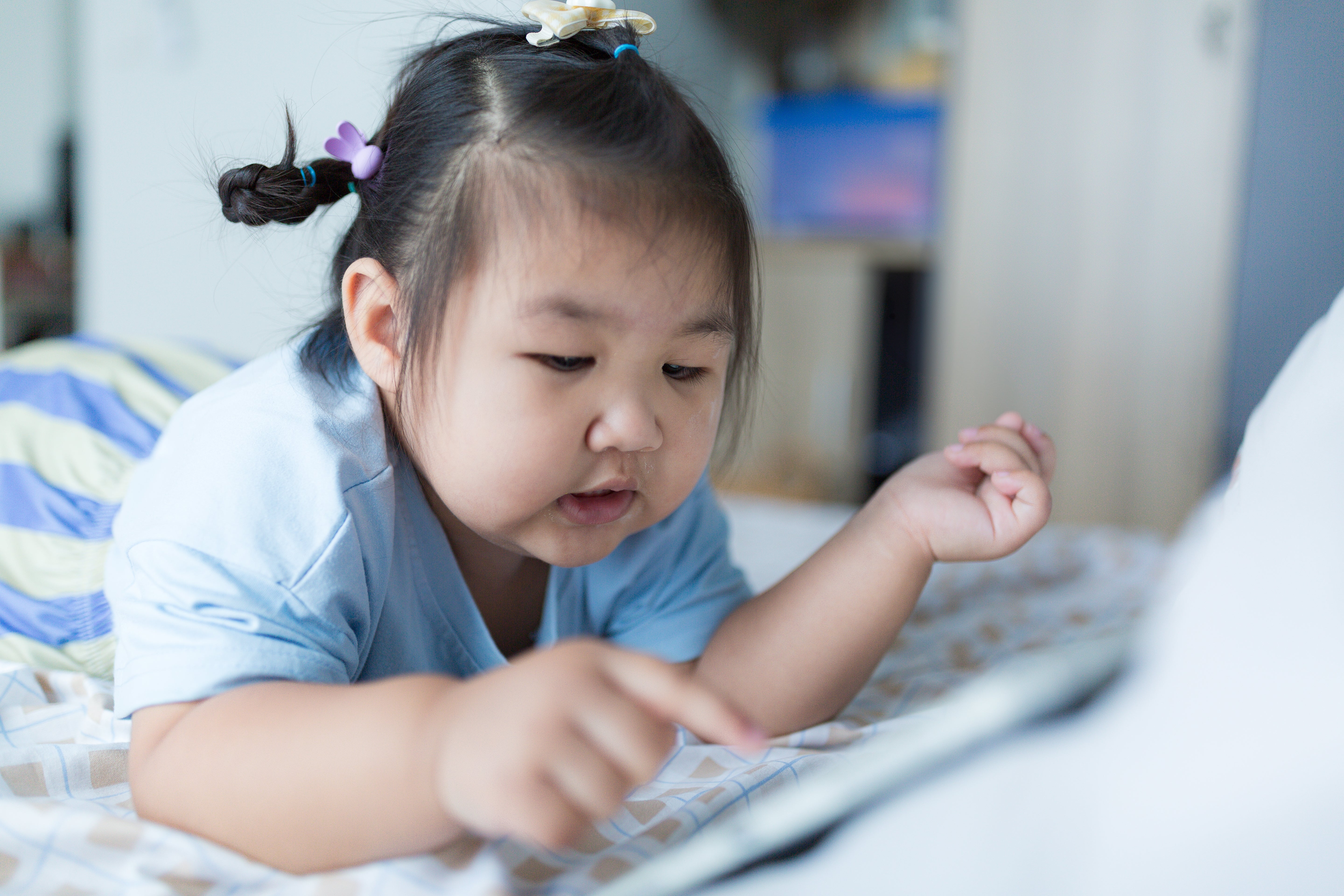 Young child lying on a bed, using a tablet. She is focused and engaged, with hair tied up in playful ponytails secured with clips