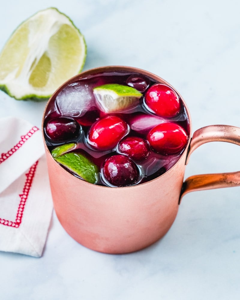 Mule mug filled with iced cranberry cocktail, garnished with lime wedges and cranberries, adjacent  to a cloth napkin and lime wedge