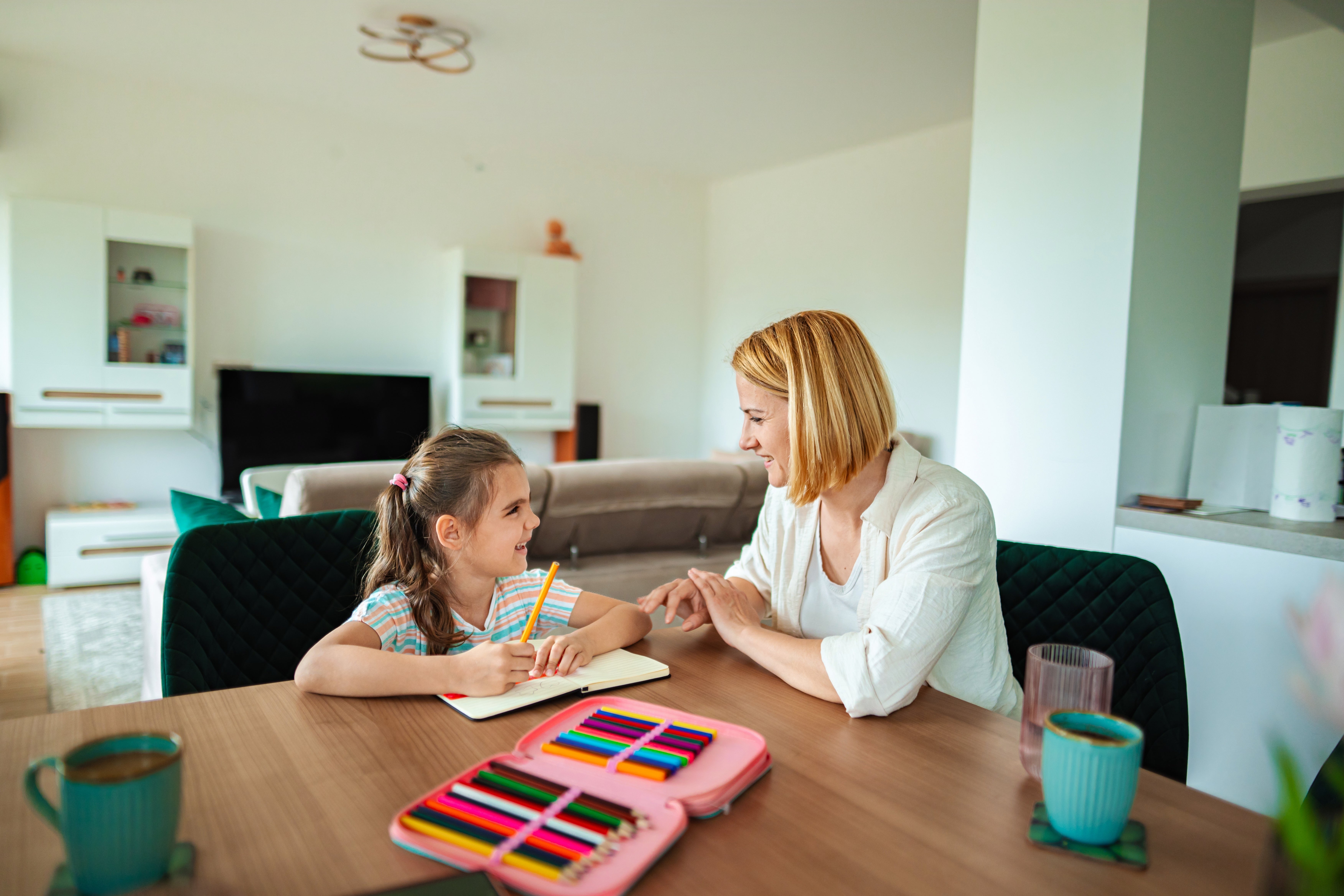Mother and daughter sitting at a table, smiling while doing homework together. Table has colored pencils and cups. Cozy living room in the background