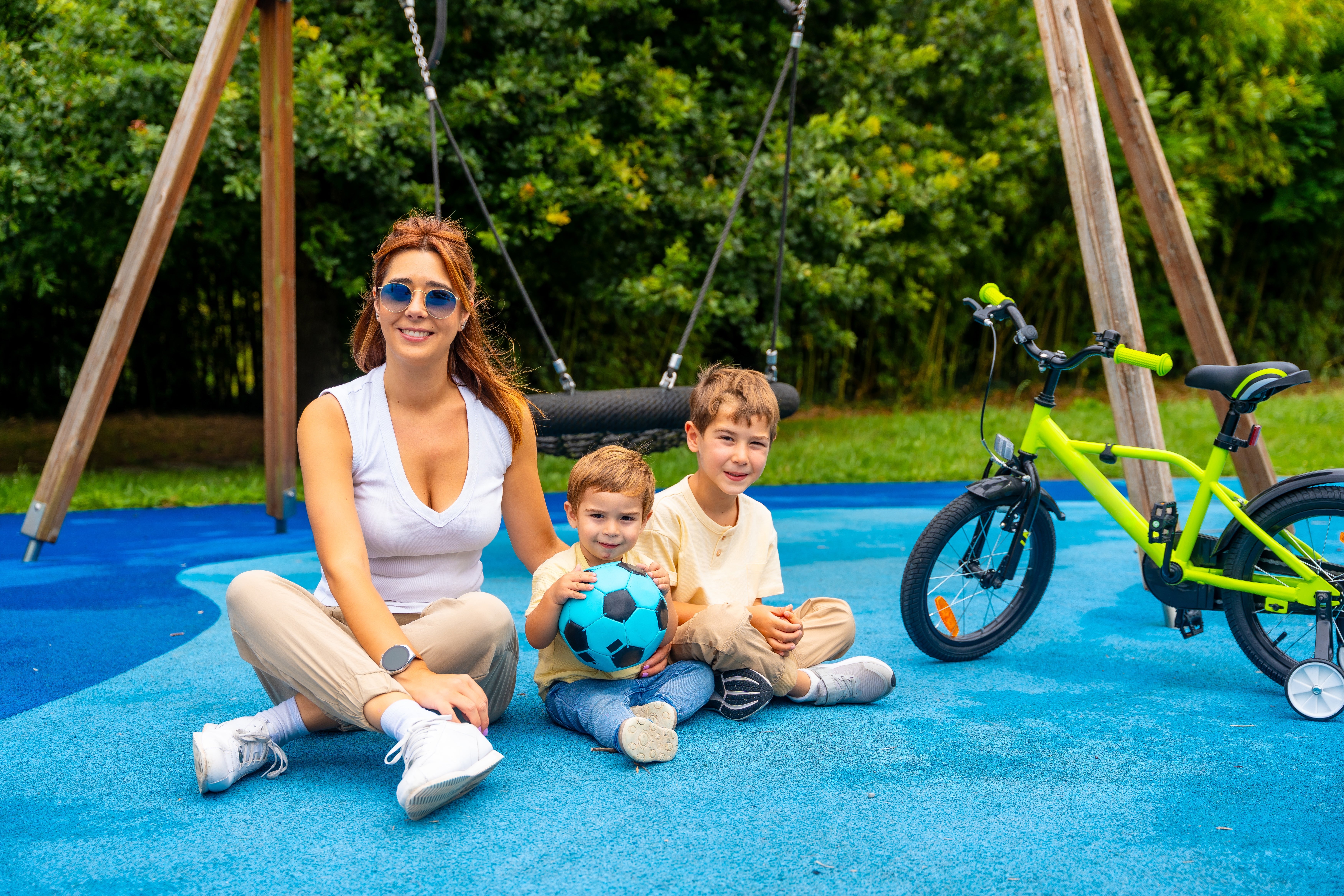 A mother and her two young children sit on a playground, smiling and holding a soccer ball, with a bicycle nearby