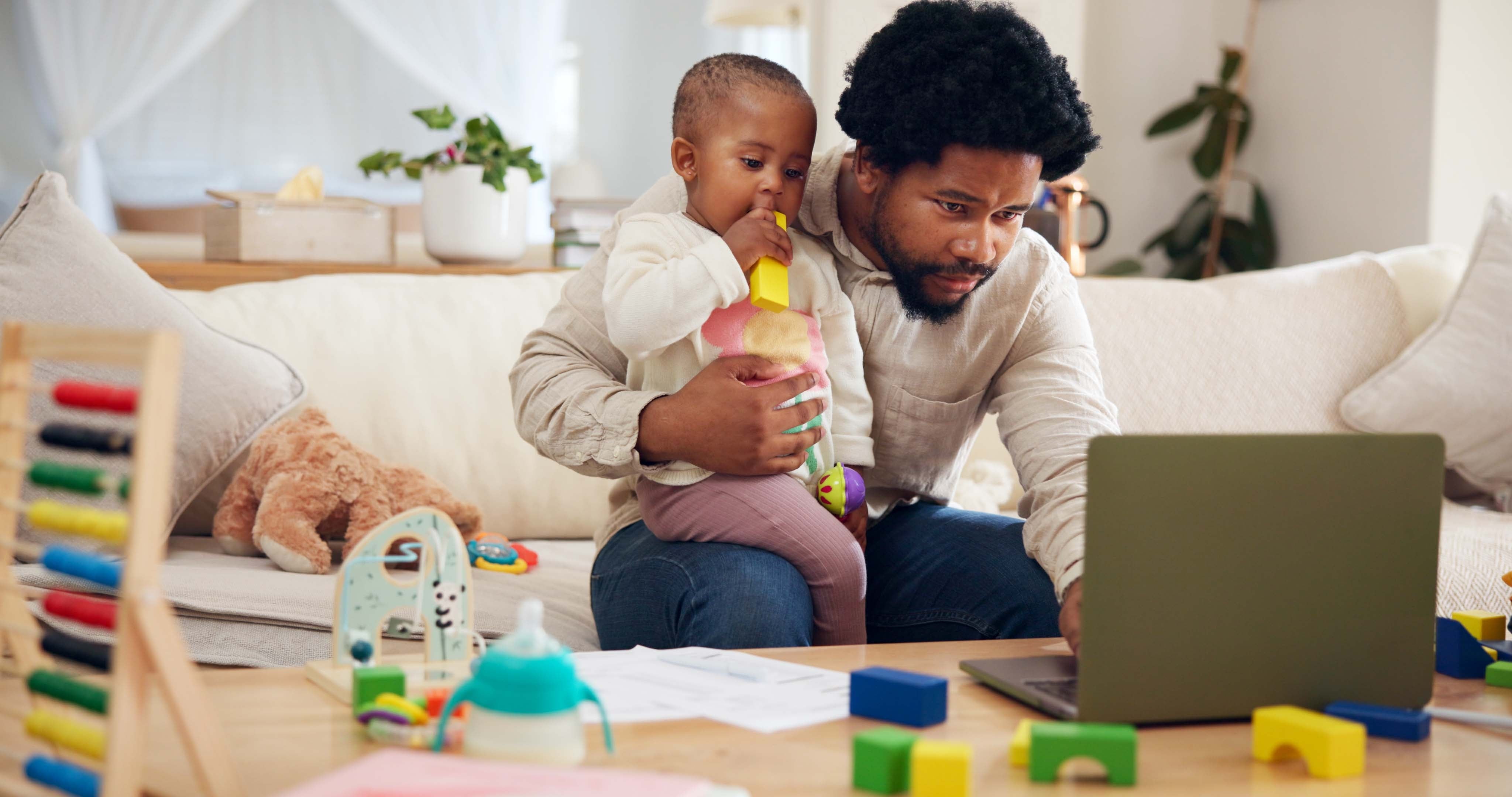 Father working on a laptop while holding a young child who is playing with a toy in a cozy living room with scattered toys