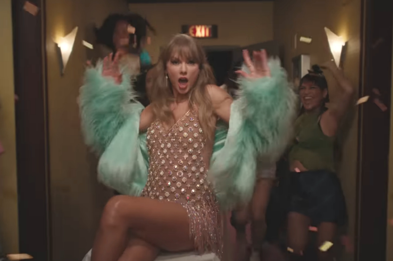 Singer in a glittery dress and feathered jacket surrounded by dancing people in a lively hallway scene