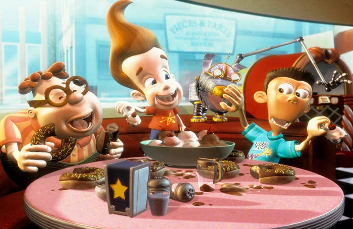 Three animated characters, including one with tall, swooping hair, excitedly examine a robotic object in a retro-style diner with scattered snacks
