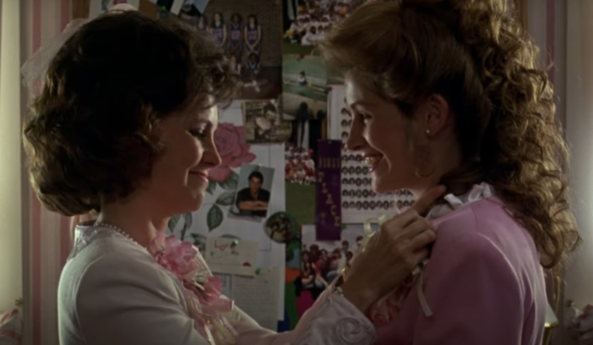 Two women smiling astatine  each   other, adjusting a corsage. They are successful  a country   with a collage of photos connected  the partition  down  them