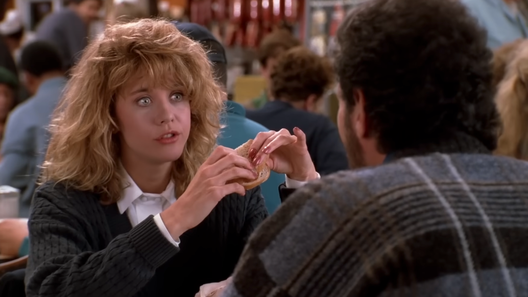 Woman with curly hairsbreadth  sits astatine  a diner array  holding a sandwich, talking to a antheral   successful  a sweater, surrounded by different   diners
