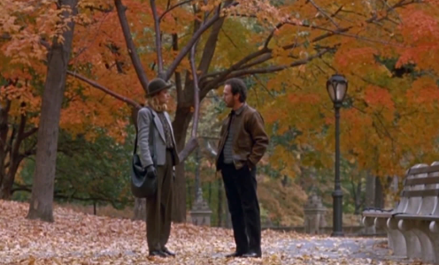 Two radical   basal   talking connected  a leaf-covered way  successful  a parkland  during autumn, surrounded by trees with autumn  foliage. One idiosyncratic   wears a chapeau  and coat