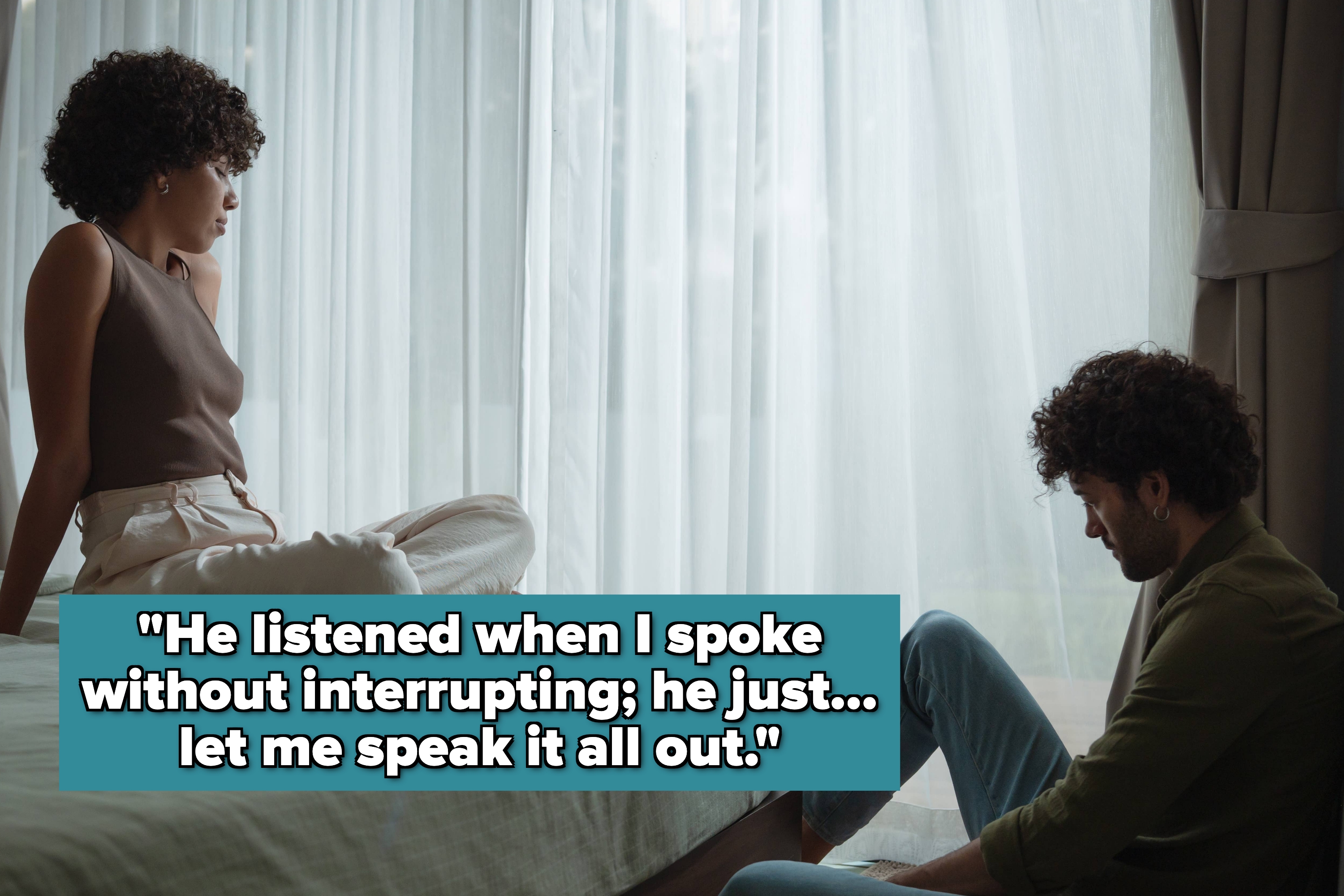 A woman sits on a bed looking at a man sitting on the floor, both with thoughtful expressions in a softly lit room