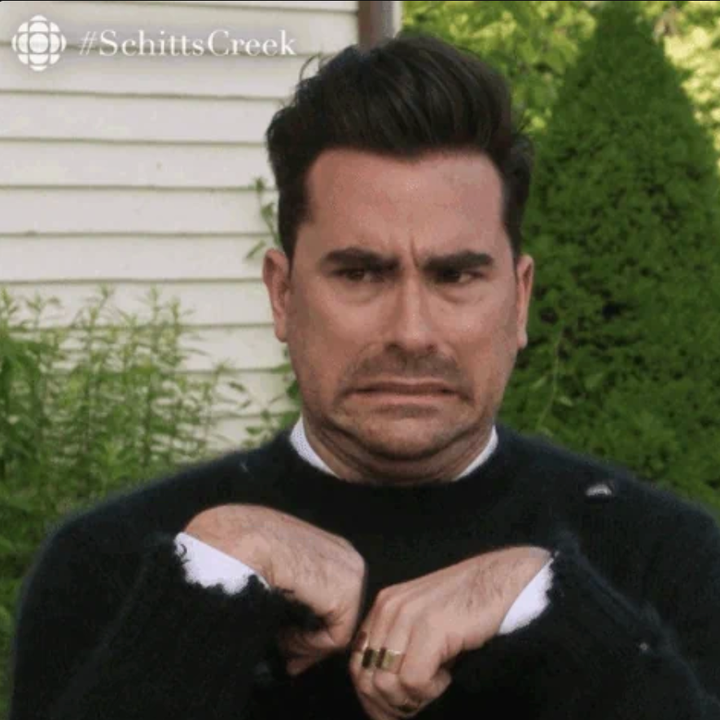 A person with a grimacing expression and raised hands mimics a nervous gesture. Background includes a house exterior and greenery. #SchittsCreek logo