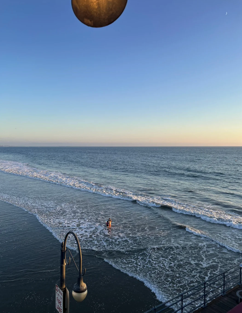 A idiosyncratic   stands astatine  the shoreline connected  a calm formation  during sunset, with gentle waves approaching. A streetlamp is partially disposable   successful  the foreground