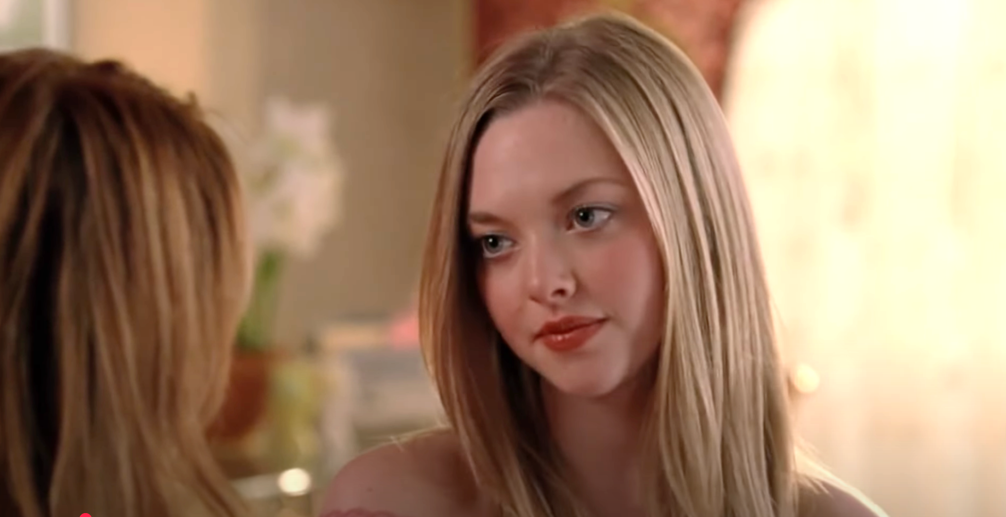 Two people are in conversation indoors, facing each other closely. Both have long hair and are dressed casually