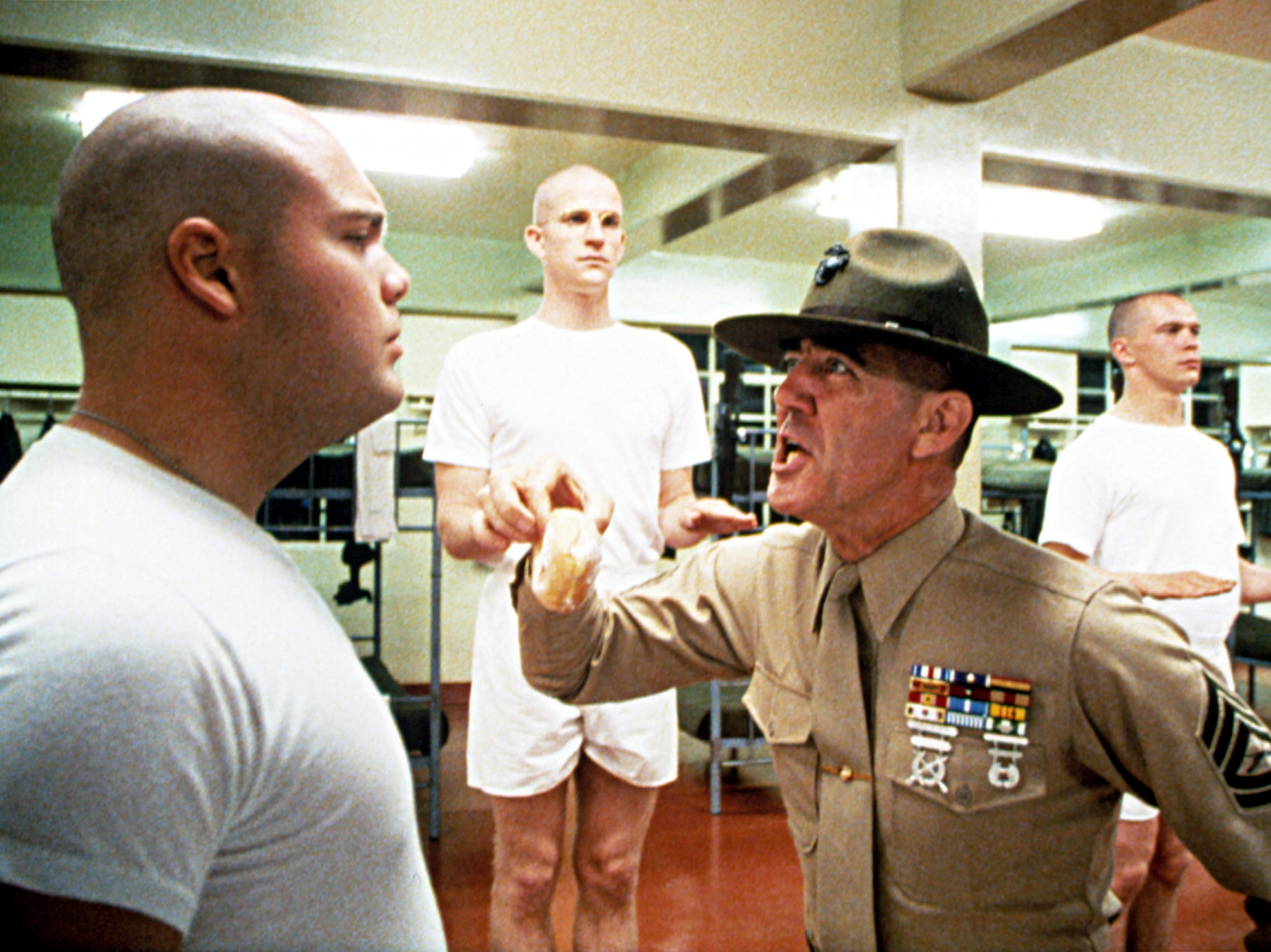 Ermey as a drill instructor in uniform yells at a recruit in a military barracks scene