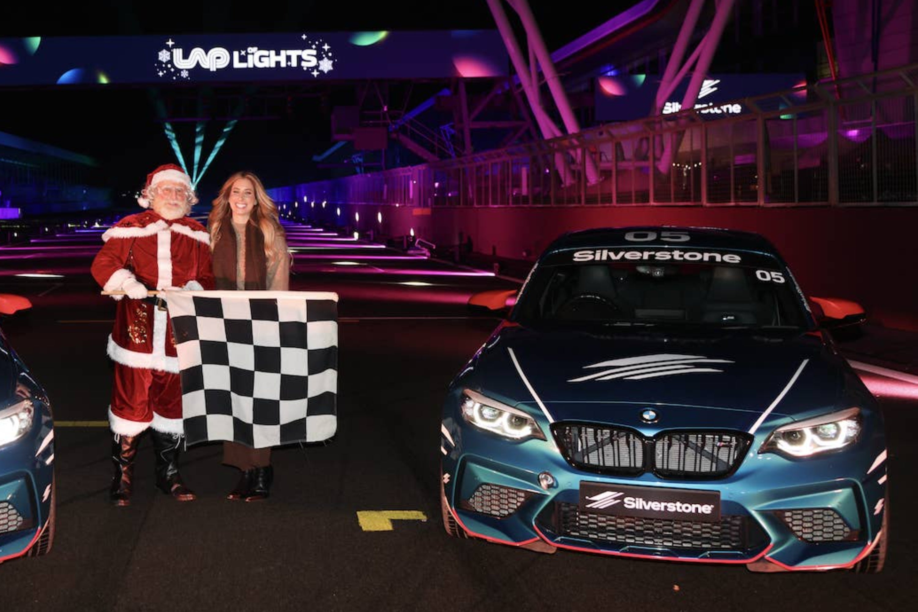 Person in a Christmas sweater and Santa hat poses joyfully on a podium. Christmas-themed display reads “Silverstone Lap of Lights.”