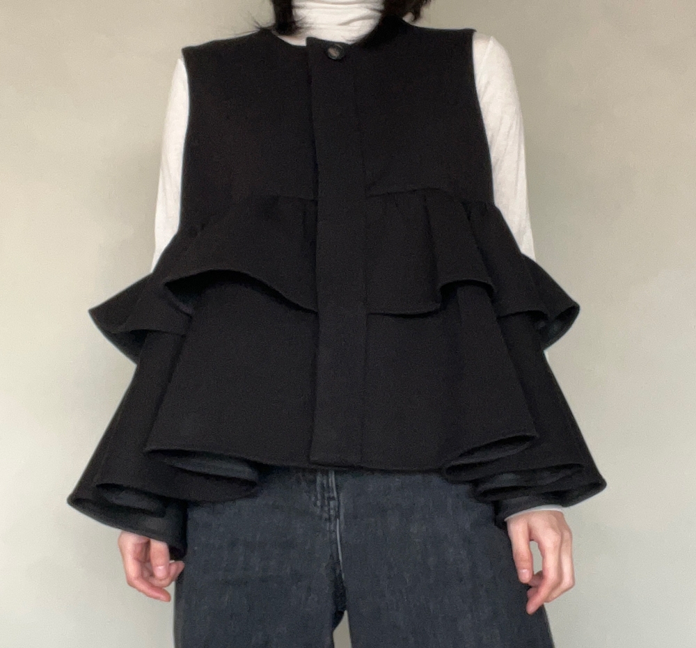 Person wearing a layered, flared black top over a white turtleneck, paired with dark pants, against a plain background