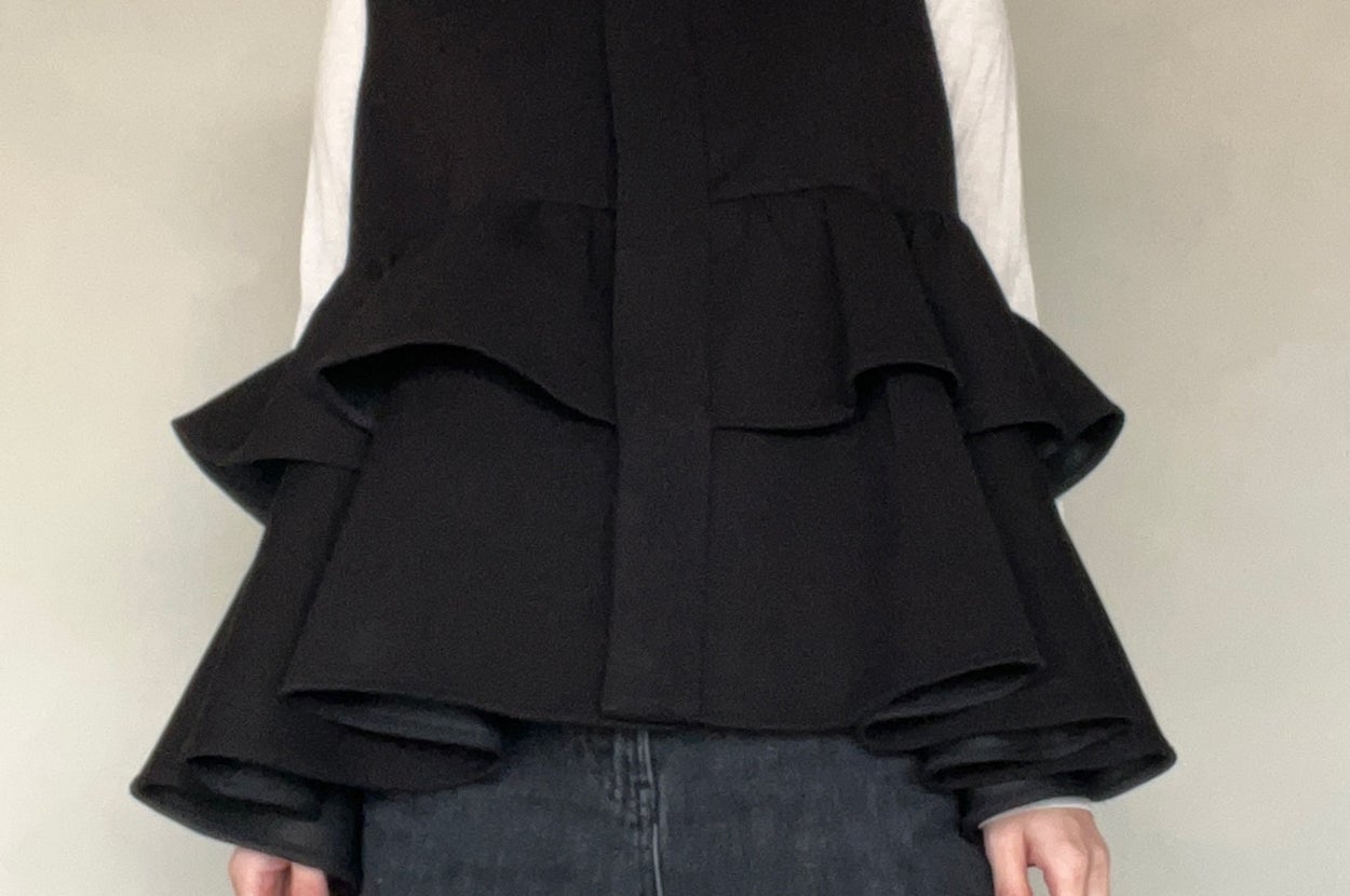 Person wearing a layered, flared black top over a white turtleneck, paired with dark pants, against a plain background