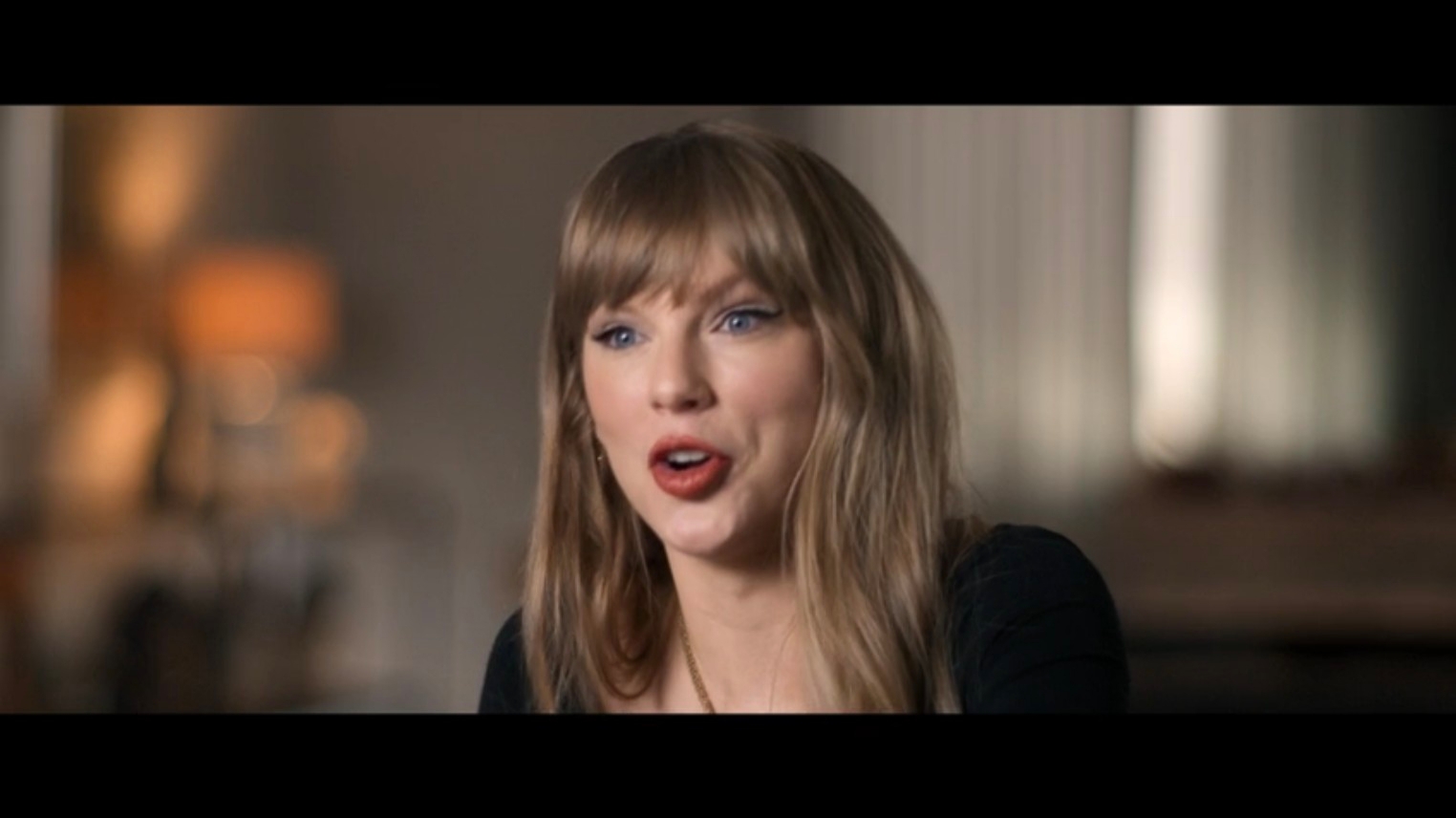 A idiosyncratic   with agelong  hair, wearing a casual top, speaks animatedly indoors
