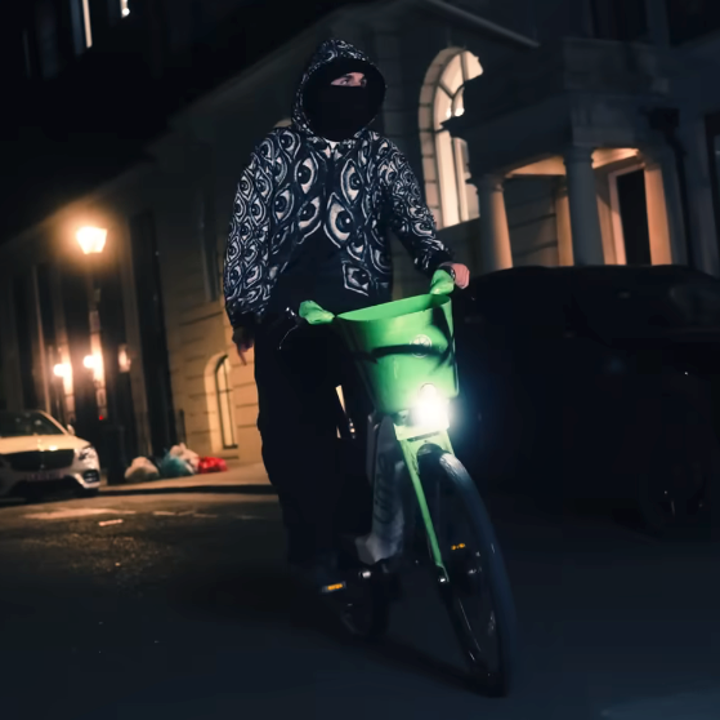 Person in patterned hoodie and face mask rides a bicycle at night on a city street