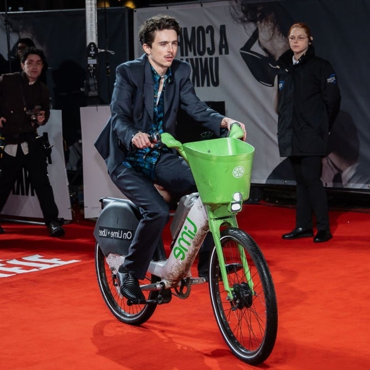 Person in formal suit rides a green rental bike on a red carpet, with photographers and attendees in the background