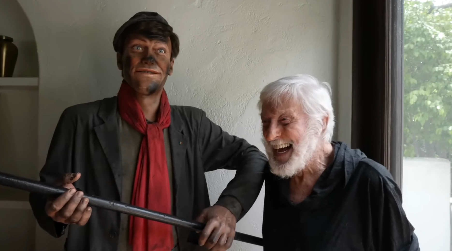 An aged  antheral   laughs adjacent  to a statue of a quality  wearing a cap, reddish  scarf, and holding a chimney expanse  brush