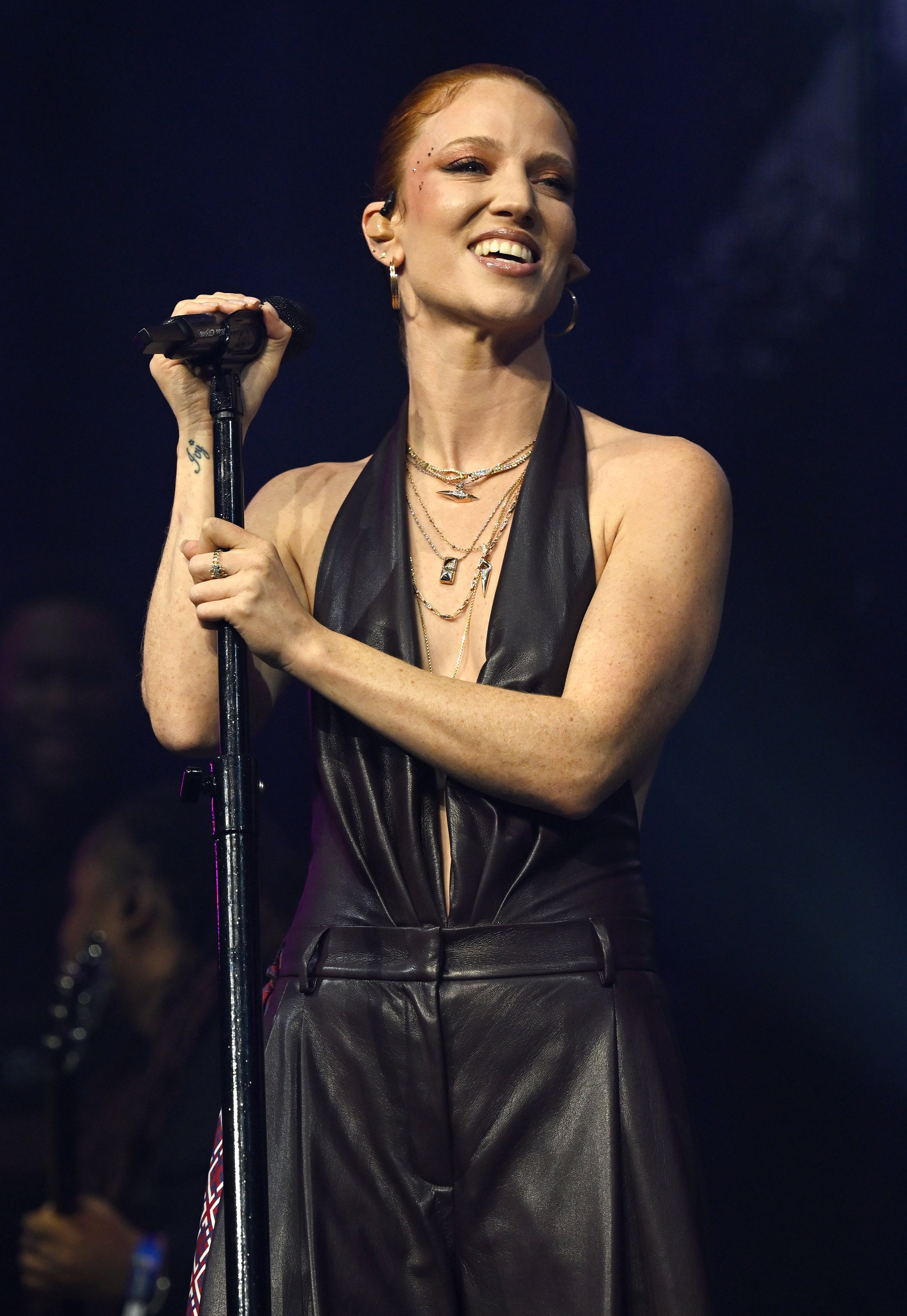 Sanger som opptrer på scenen, smiler, iført et ermeløst skinnantrekk med lagdelte halskjeder