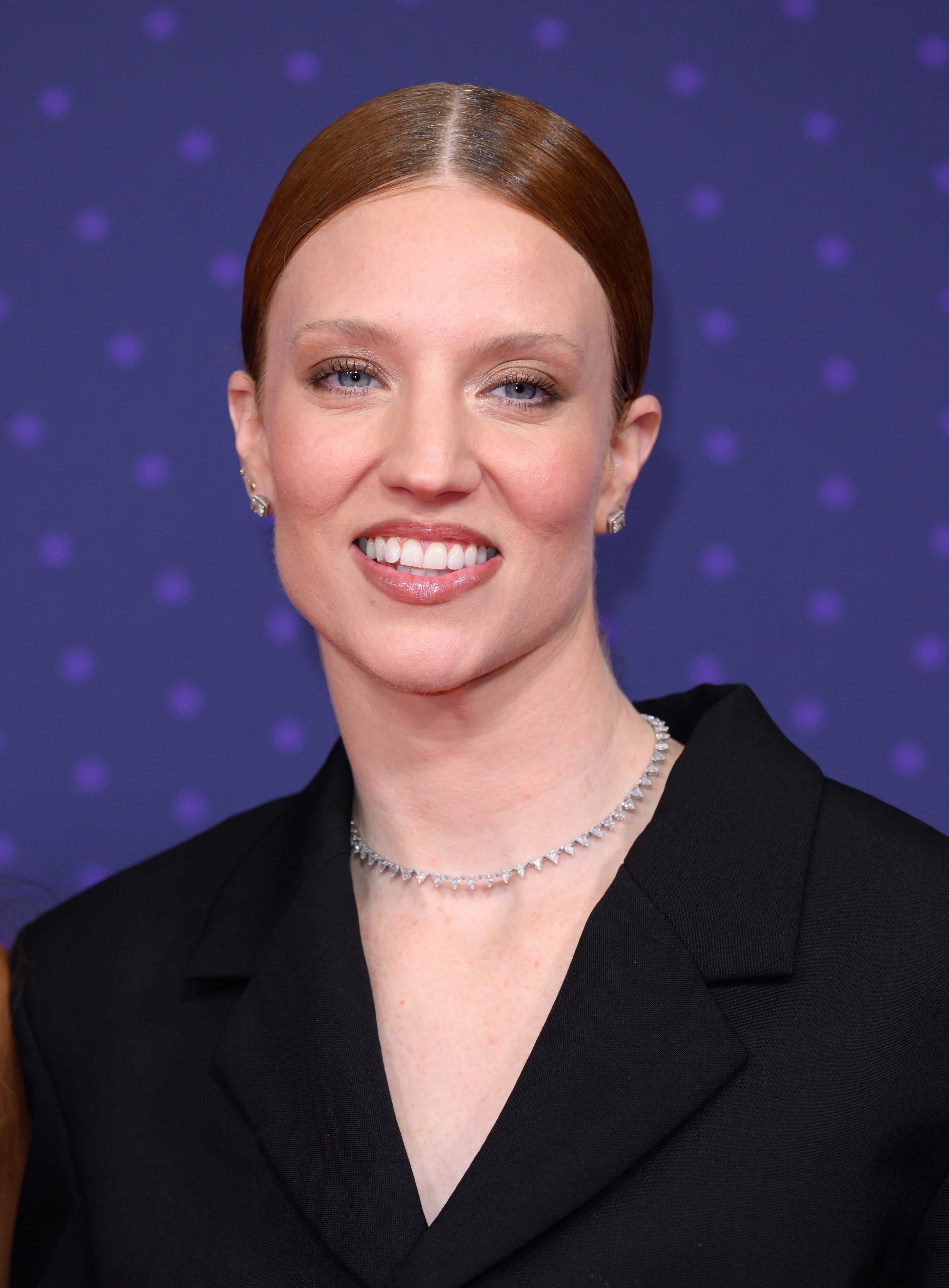 Person som smiler på et arrangement, iført en mørk blazer og diamantkjede, med hår stylet i en elegant updo mot et prikkete bakteppe