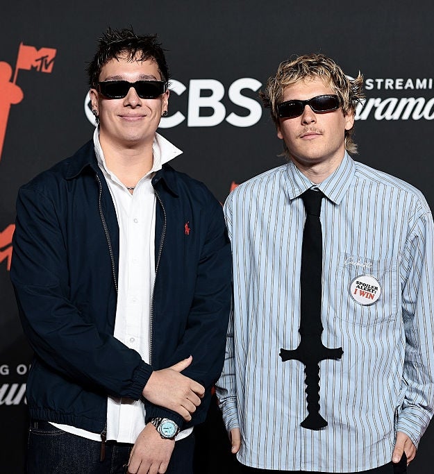 To personer på en rød løper iført uformelle antrekk og solbriller på et MTV-arrangement i bakgrunnen