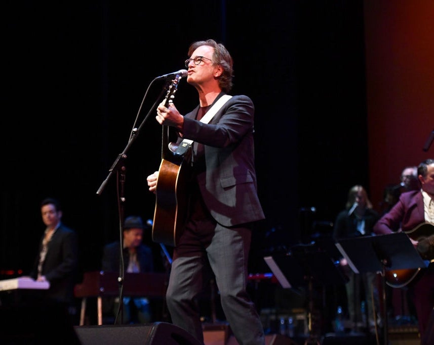 Musiker opptrer på scenen med gitar, iført mørk dress og briller. Keyboardist og andre bandmedlemmer er i bakgrunnen