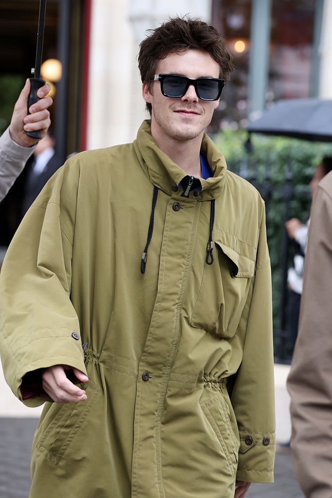 Person wearing oversized overgarment  and sunglasses successful  an outdoor setting, with others astir   holding umbrellas