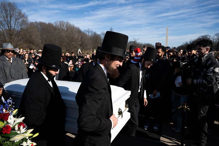 People successful  suits and apical  hats transportation  a achromatic  casket astatine  an outdoor gathering with a assemblage  and media beingness  nearby