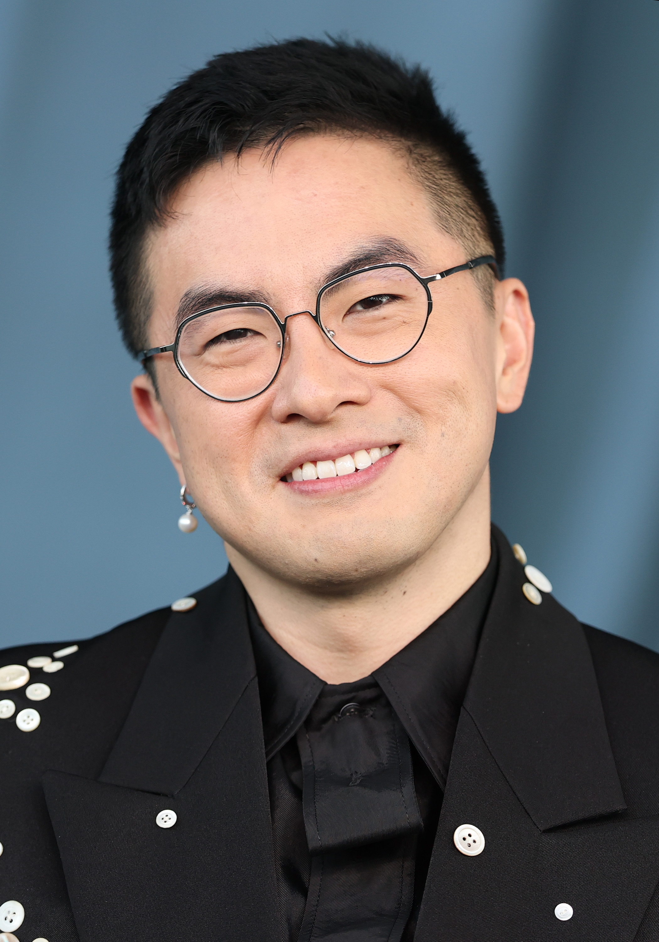 Person with abbreviated  hairsbreadth  and glasses smiles successful  a achromatic  suit   adorned with decorative pearl-like buttons