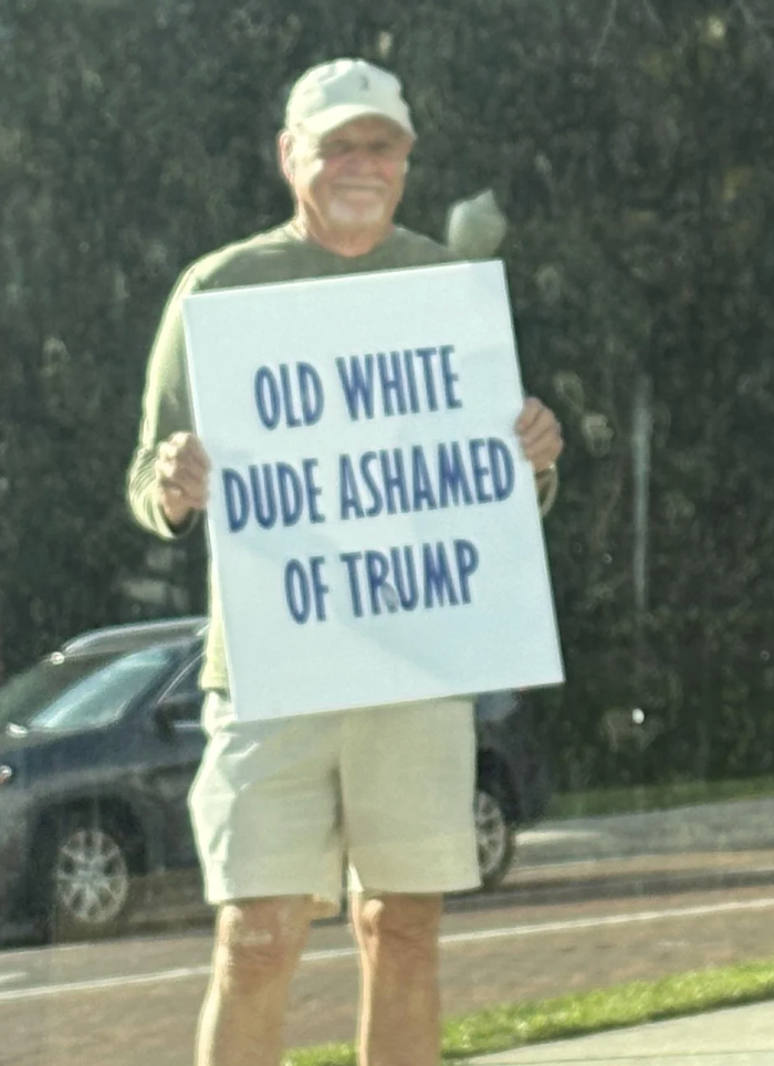 Man holding a motion   that reads, "Old achromatic  dude ashamed of Trump," lasting  connected  a sidewalk