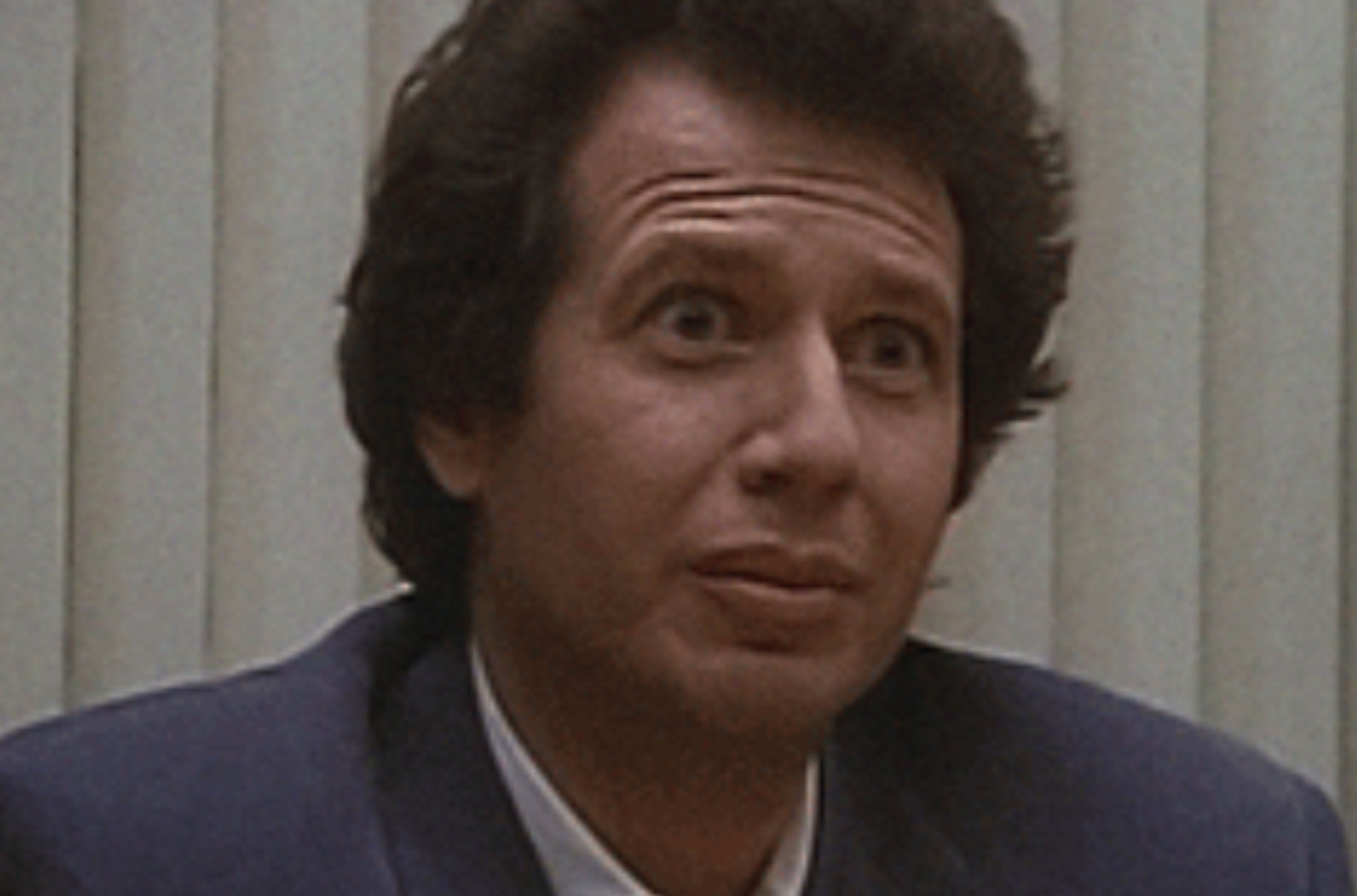 Person successful a suit, seated indoors, looking upward with a neutral expression. Professional mounting suggested