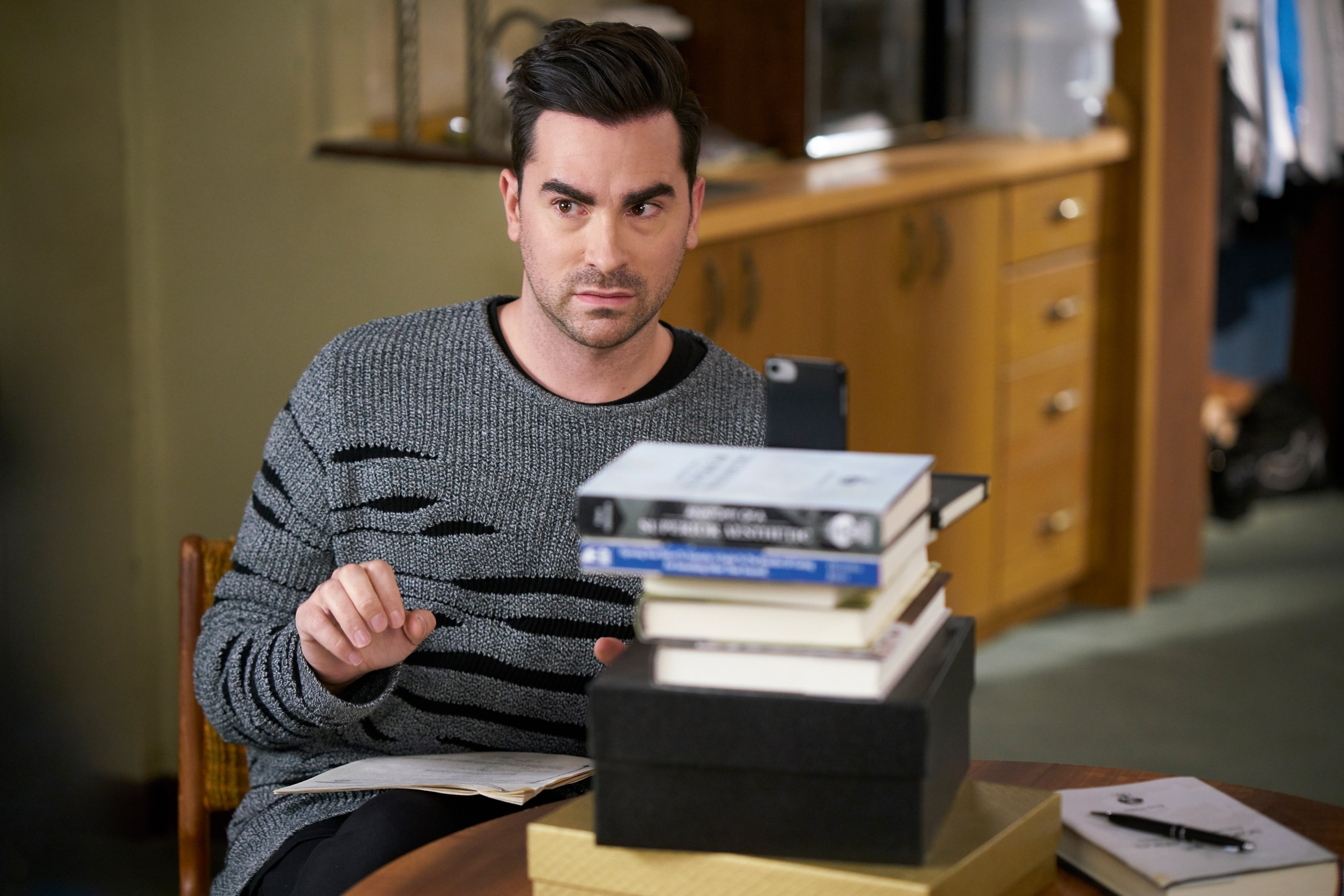 Person wearing a patterned sweater is seated astatine a array with a stack of books and a phone, looking to the broadside successful a thoughtful manner