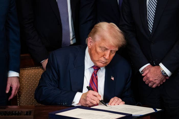 A antheral   successful  a suit   is sitting astatine  a table  signing a document, surrounded by others successful  ceremonial  attire