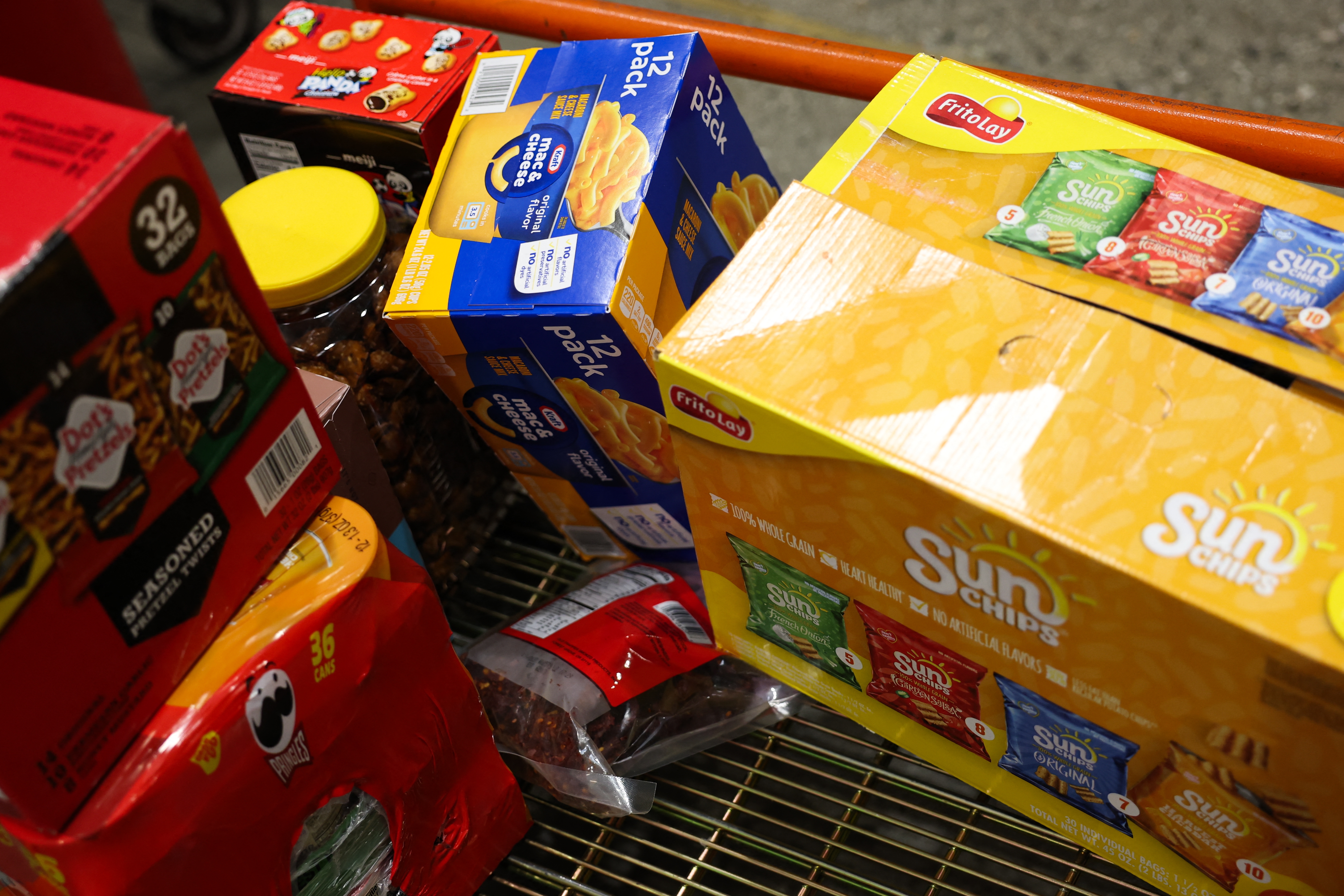 Grocery store   cart filled with snacks and packaged foods, including chips, crackers, and a jar of pretzels