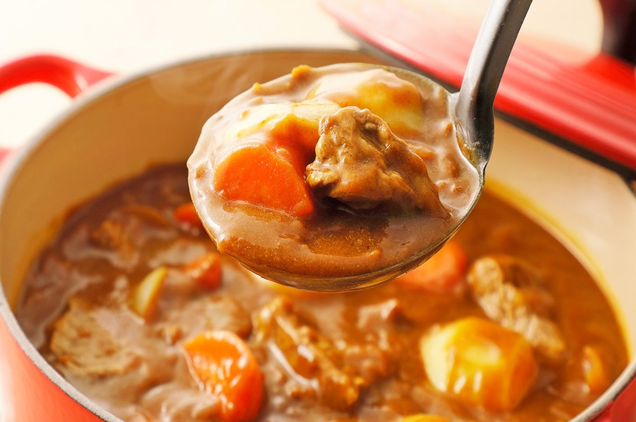 赤い鍋に入ったビーフシチューをスプーンですくう様子。野菜と肉が見える。