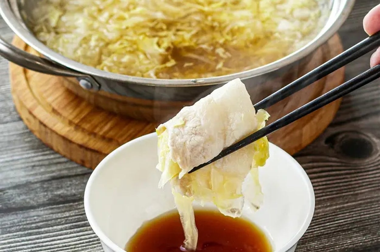 鍋料理の一部を箸で取り、タレの入った小皿に入れる様子。鍋には白菜などの野菜が入っている。