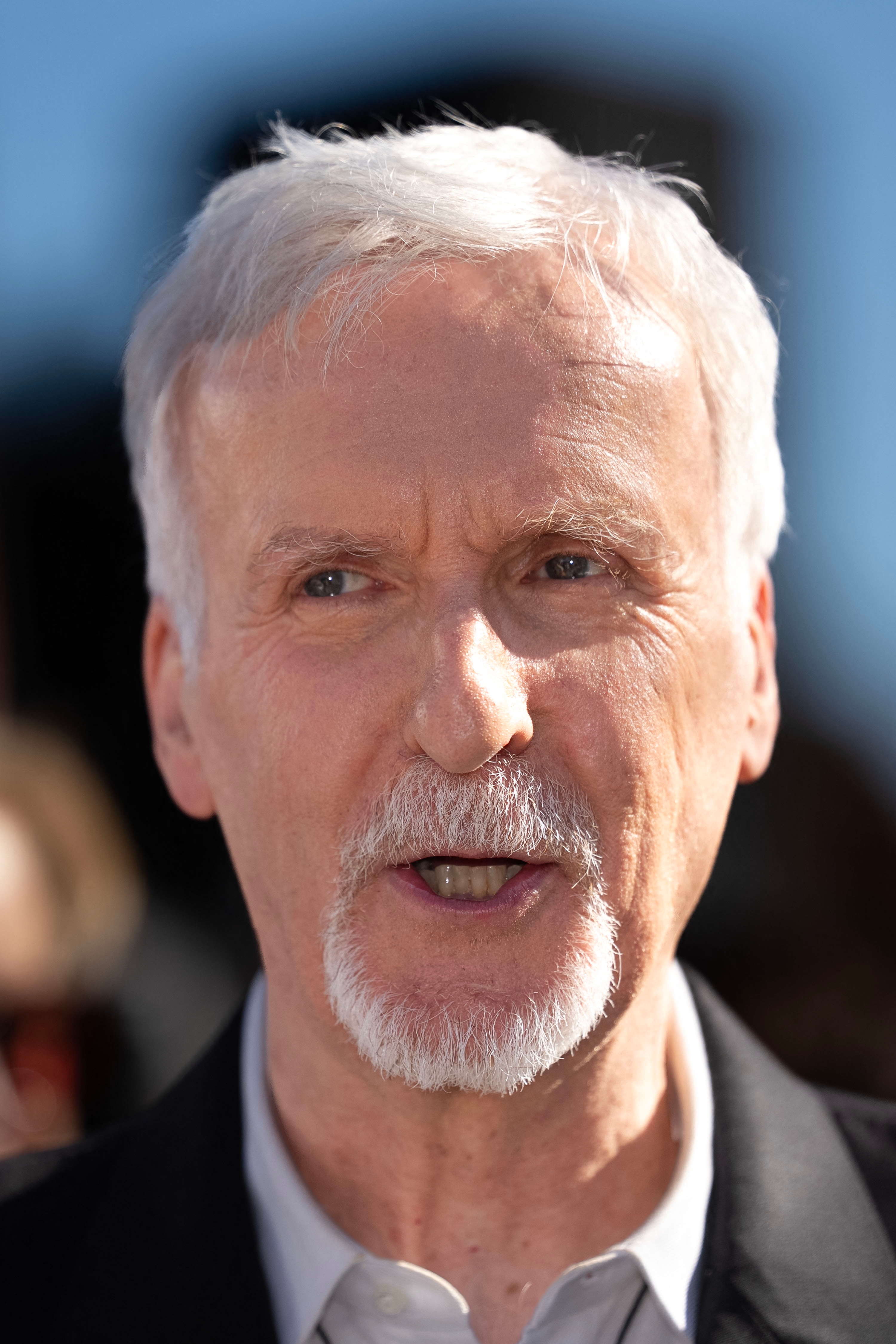 James Cameron with abbreviated  achromatic  hairsbreadth  and a goatee, wearing a collared garment  and blazer, speaks passionately outdoors