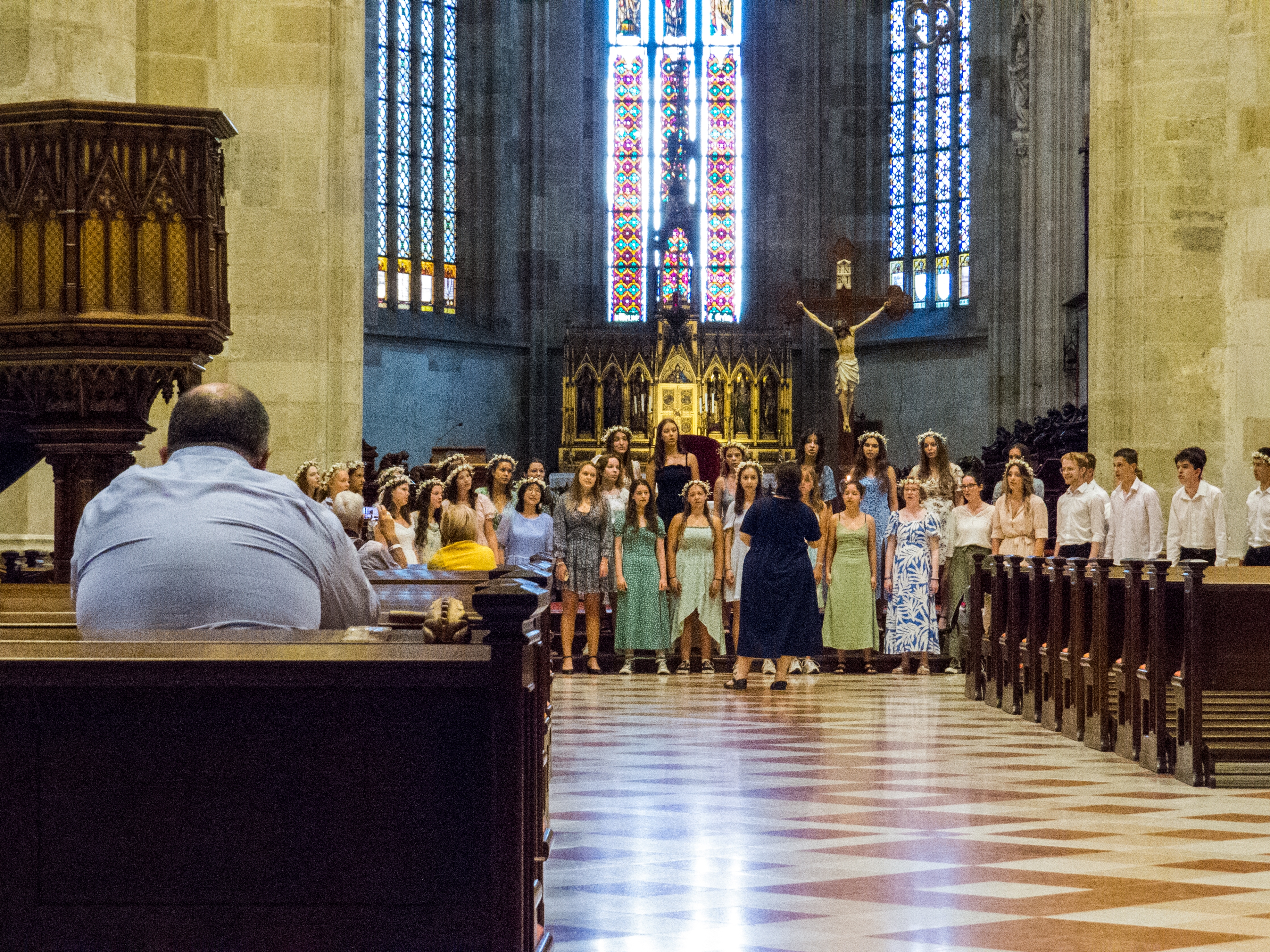 Korsang i en stor kirke, ledet av en kvinne; utsmykket alter og glassmalerier er synlige i bakgrunnen