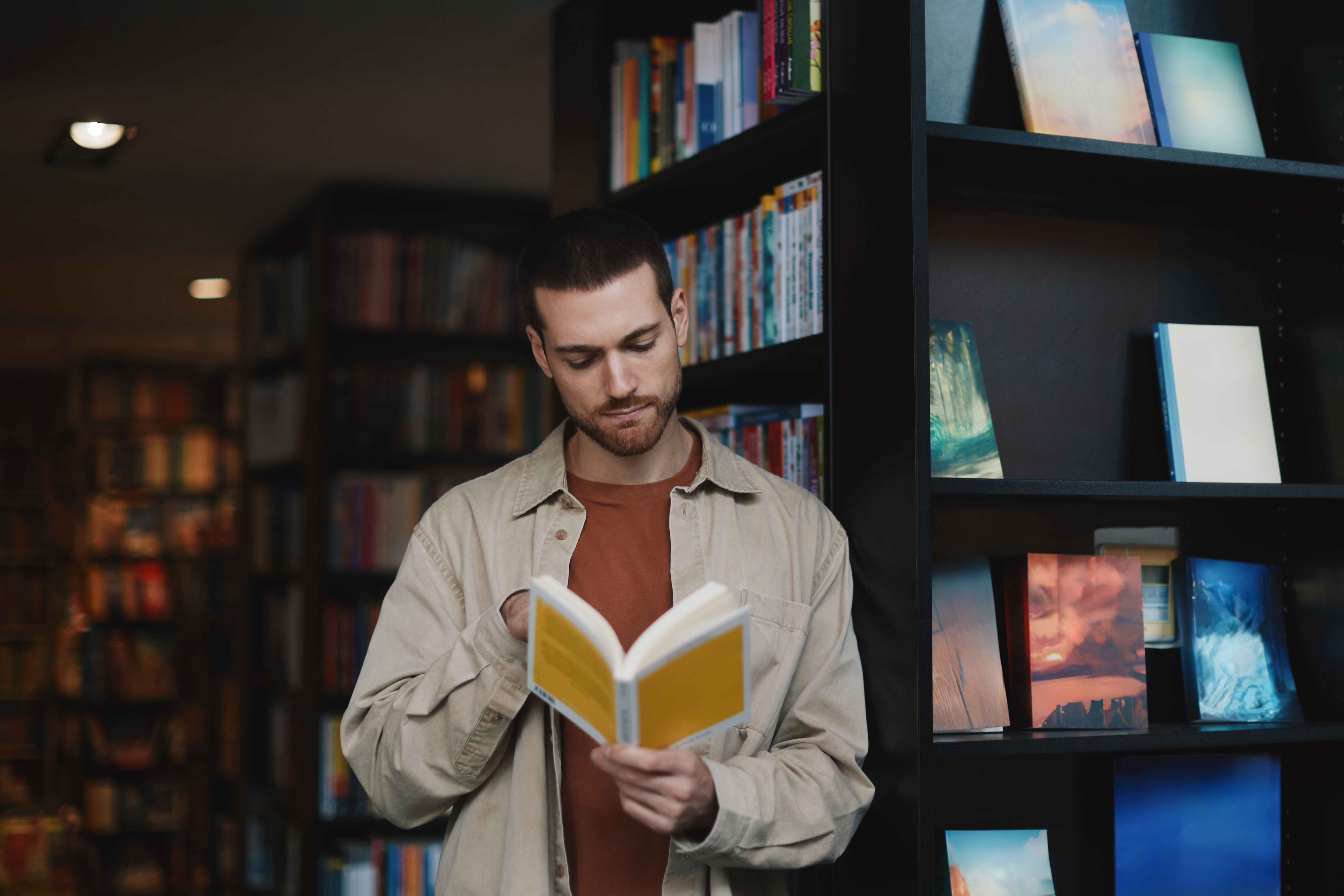 Person som leser en bok i en bokhandel, står mellom bokhyllene, fokusert og uformelt kledd