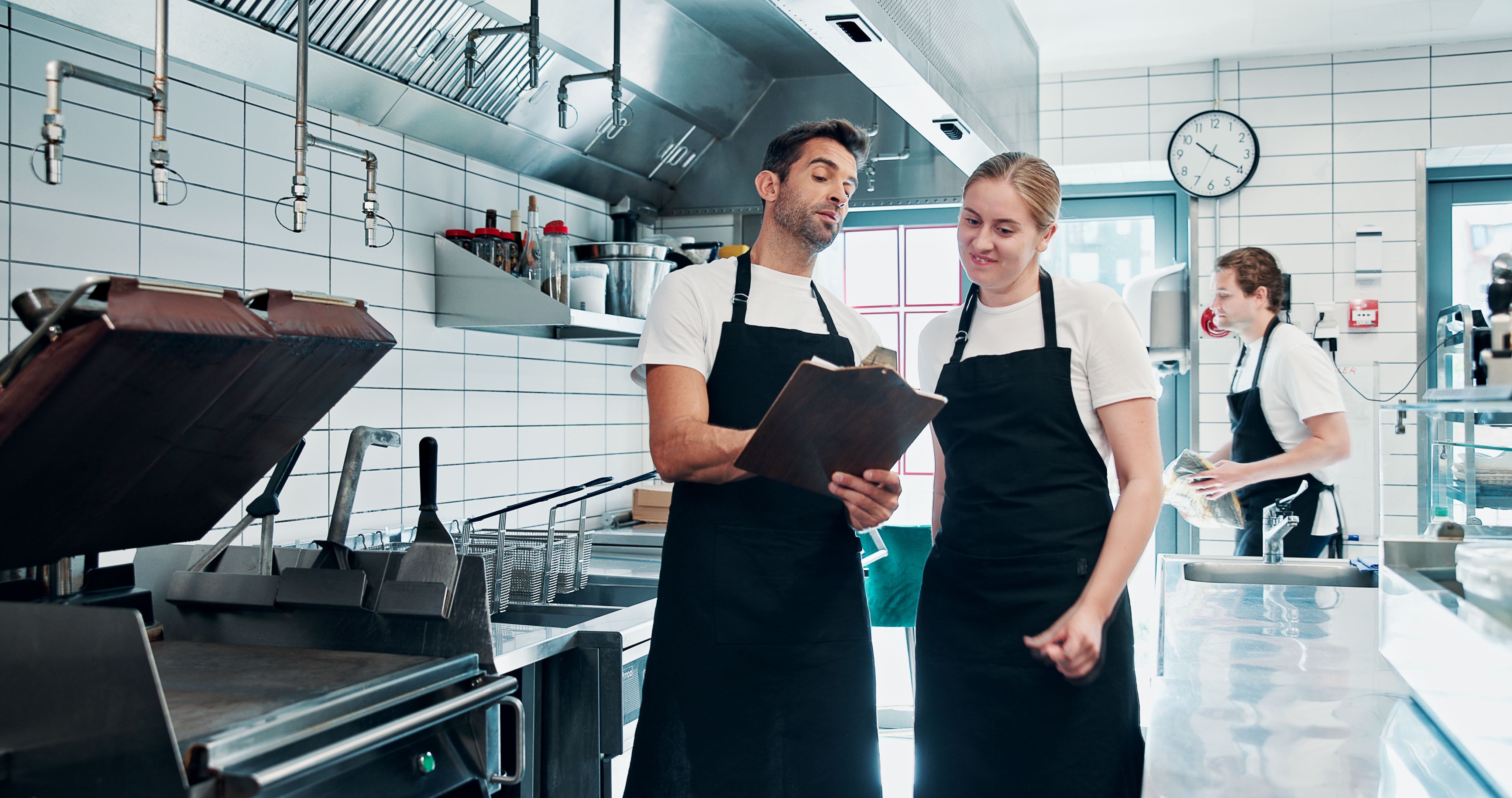 To kokker på et restaurantkjøkken diskuterer notater på en utklippstavle, iført forkle. En annen kokk jobber i bakgrunnen
