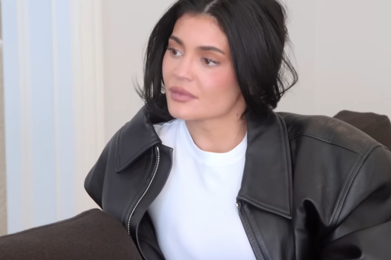 Kylie Jenner sitting on a couch, wearing a shirt and leather jacket, gazing to the side